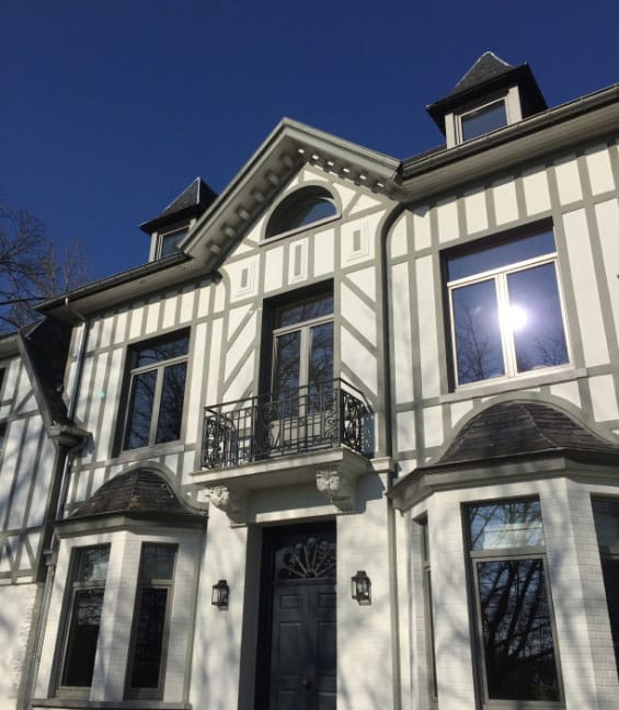 Une grande maison élégante avec une façade de style Tudor, avec des murs blancs et une charpente décorative, témoigne dune rénovation réfléchie. Elle possède de nombreuses fenêtres, un balcon central et des toits pointus. Le soleil se reflète sur une fenêtre sur un ciel bleu clair.