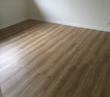 Een lege kamer met lichtgekleurde houten vloeren en witte muren, verlicht door natuurlijk licht dat door een onzichtbaar raam stroomt, biedt de perfecte setting voor een chique make-over. Stel je eens voor hoe gemakkelijk het is om clip-on vinyl over tegelvloeren te installeren om de ruimte moeiteloos te transformeren.