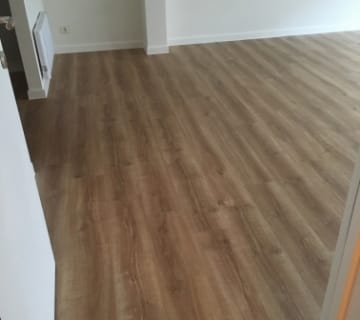 De kamer heeft een lichtbruine laminaatvloer en strakke witte muren. Een zichtbare radiator rust op de linkermuur, terwijl een gedeeltelijk open deur op de voorgrond te zien is. Stel je voor dat je deze ruimte transformeert met clip-on vinyl over tegelvloer voor een moeiteloos chique upgrade.