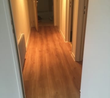 Een gang met clip-on vinyl over houten vloer en witte muren, met een open deur aan het einde. De ruimte is verlicht met warme, sfeervolle verlichting.