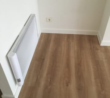 Een kleine aan de muur gemonteerde kachel staat in een kamer met een lichte houten vloer, die lijkt op vinylclips, en ongerepte witte muren. Een stopcontact is zichtbaar boven de vloer. De lege hoek van de kamer benadrukt de contrasterende texturen tussen de vloer en de muren.