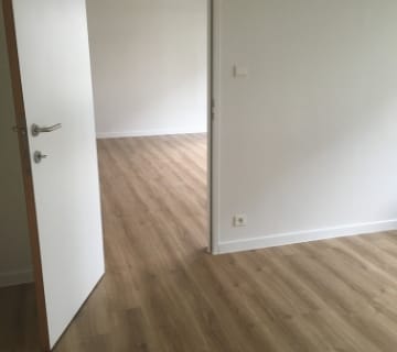Een minimalistisch interieur met een lichte houten vloer en eenvoudige witte muren, met een open deur die twee kamers met elkaar verbindt. Clip-on vinyl op een tegelvloer benadrukt de eenvoud en vergroot de ruimtelijkheid.