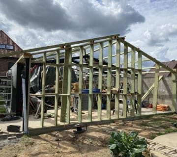 La structure en bois dun bâtiment, partiellement construite, se trouve à lextérieur sous un ciel nuageux. Divers matériaux de construction et outils sont éparpillés un peu partout, laissant deviner le travail dun habile toiturier. Au premier plan, quelques plantes poussent dans un jardin.