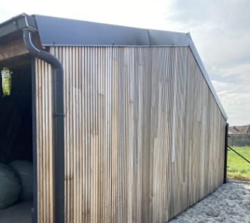 Une grange moderne avec des lattes verticales en bois clair et un toit en métal foncé réalisé par des couvreurs experts, située dans un champ. La grange dispose dun tuyau de descente sur le côté et de balles de foin à lintérieur, visibles à travers le côté ouvert. Le ciel est couvert.