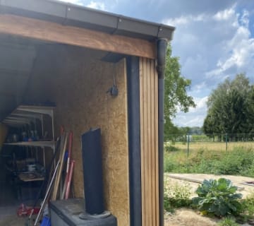 Une porte de garage partiellement ouverte révèle des étagères avec des outils, laissant deviner le travail dun toiturier à lintérieur. À lextérieur, le ciel est partiellement nuageux et un jardin avec une grande plante feuillue et des arbres est visible, entouré dune clôture.