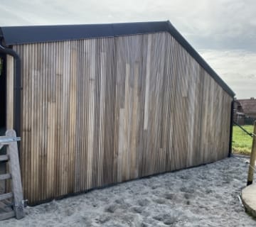 Un bâtiment moderne en bois, aux panneaux verticaux et au toit en pente, réalisé par des couvreurs qualifiés, se dresse sur un sol sablonneux. En arrière-plan, de petites zones clôturées sont visibles sous un ciel clair parsemé de nuages.