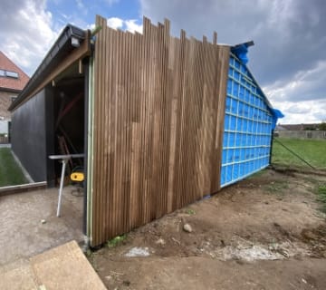 Un petit bâtiment en construction présente un mur partiellement recouvert de lattes de bois verticales, habilement installées par un toiturier. Le côté opposé présente des panneaux isolants bleus apparents. Une scie est visible à gauche et le ciel au-dessus est partiellement nuageux.