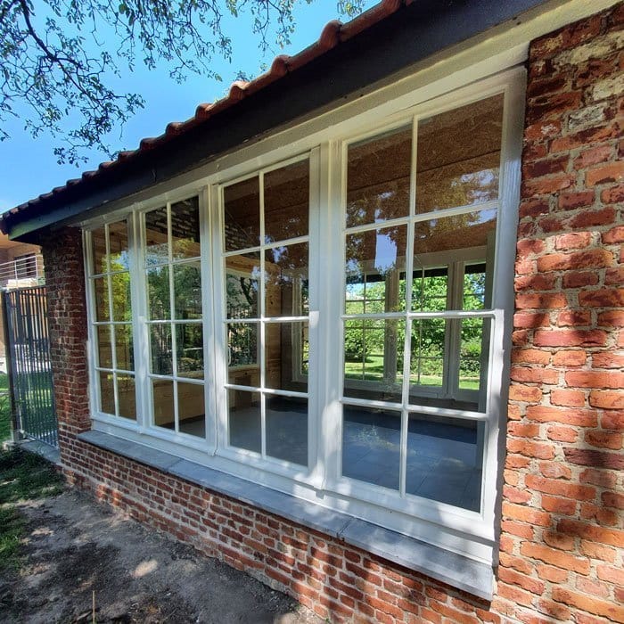 Un bâtiment en brique avec de grandes fenêtres vitrées reflétant la verdure environnante met en valeur le talent artistique dun peintre en bâtiment qualifié. Lintérieur semble vide, avec un sol carrelé, tandis que la lumière du soleil filtre à travers les arbres, projetant des motifs dombres complexes sur les vitres.