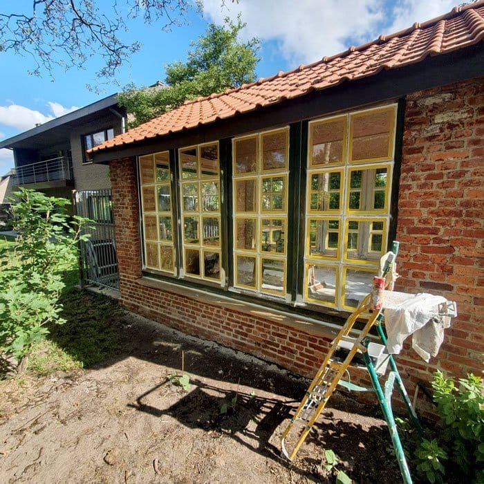 Une maison en briques rouges avec un toit en tuiles est en cours de rénovation par un peintre en bâtiment qualifié. Un échafaudage et une échelle avec un pot de peinture sont installés à côté de fenêtres aux cadres jaunes fraîchement peints. Le bâtiment est entouré de verdure sous un ciel partiellement nuageux.