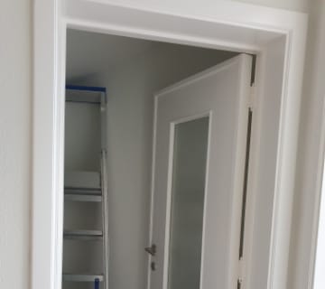 Une porte blanche partiellement ouverte avec une vitre est encastrée dans un mur blanc. Derrière elle, une échelle métallique en forme descalier sappuie contre le mur à lintérieur de la pièce, ajoutant une touche de charme industriel à lespace intérieur épuré et minimaliste.