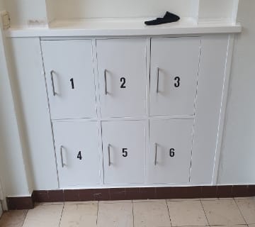 Un ensemble de six casiers blancs, numérotés de 1 à 6, est encastré dans un mur à côté de lescalier. Chaque casier est doté dune poignée en métal. Une seule chaussette noire repose sur létagère au-dessus des casiers. Le sol est carrelé de carreaux beiges et marron.