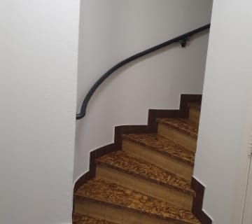 Un escalier en colimaçon aux marches marron et à la rampe noire épurée sélève entre des murs blancs immaculés. Les marches texturées suggèrent une élégance subtile. La scène se déroule à lintérieur, laissant entrevoir le charme dun intérieur résidentiel ou dun petit immeuble confortable.
