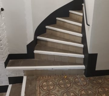 Un escalier étroit et courbé aux marches marron et aux bordures blanches mène vers le haut. Une rampe noire suit le contour, faisant écho à sa tournure élégante. Le sol à la base présente un motif de carrelage décoratif aux tons terreux.