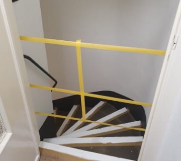 Un élégant escalier aux marches alternées de noir et de blanc est partiellement barré par une barrière horizontale jaune. Les murs environnants restent blancs, contrastant avec la robuste rampe métallique visible à gauche.