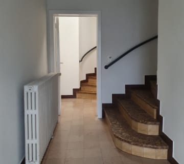 Un couloir au sol carrelé beige mène à un escalier incurvé sur la droite avec une rampe noire. Un radiateur blanc est posé sur le mur de gauche et une porte ouverte révèle une zone bien éclairée en arrière-plan.