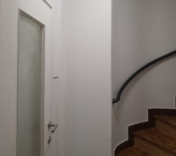 Un couloir blanc avec une porte à panneau de verre texturé à gauche et un élégant escalier avec une rampe noire à droite. Lescalier est doté de marches marron et les murs sont peints en blanc.