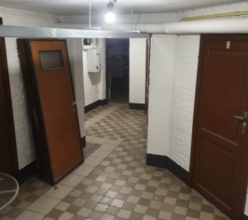 Un couloir de sous-sol faiblement éclairé avec un sol carrelé, des murs en briques blanches et plusieurs portes marron. Une porte en bois est partiellement ouverte sur la gauche, révélant un escalier. Une roue de vélo est visible dans le coin inférieur gauche. Un éclairage zénithal et des tuyaux courent le long du plafond.