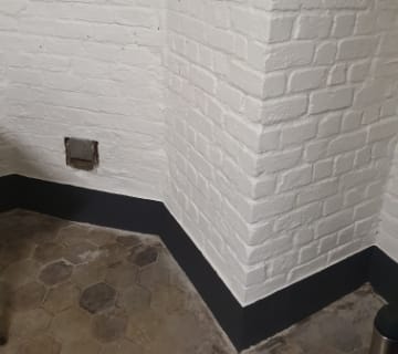 Un coin pittoresque dune pièce avec des murs en briques peints en blanc et un sol en carrelage hexagonal, rappelant le charme dun escalier. Une petite ventilation orne un mur, tandis que la base est peinte en noir, ajoutant du contraste à la scène.