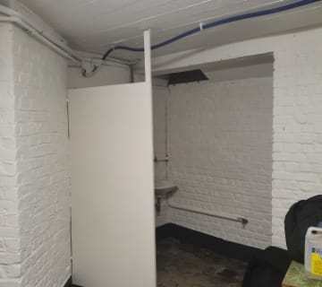 Une petite pièce en briques blanches avec une porte partiellement ouverte mène à un escalier. À lintérieur, un évier est fixé au mur avec des tuyaux visibles au-dessus. Le sol est fait de carreaux de pierre texturés et il y a un sac à dos noir sur une table.