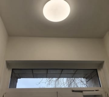 Un plafonnier rond illumine lescalier, projetant une douce lumière sur la zone. En dessous, une fenêtre rectangulaire révèle une vue sur le ciel nuageux et les branches des arbres. Le cadre blanc de la fenêtre sharmonise parfaitement avec les murs et le plafond, créant une esthétique harmonieuse.