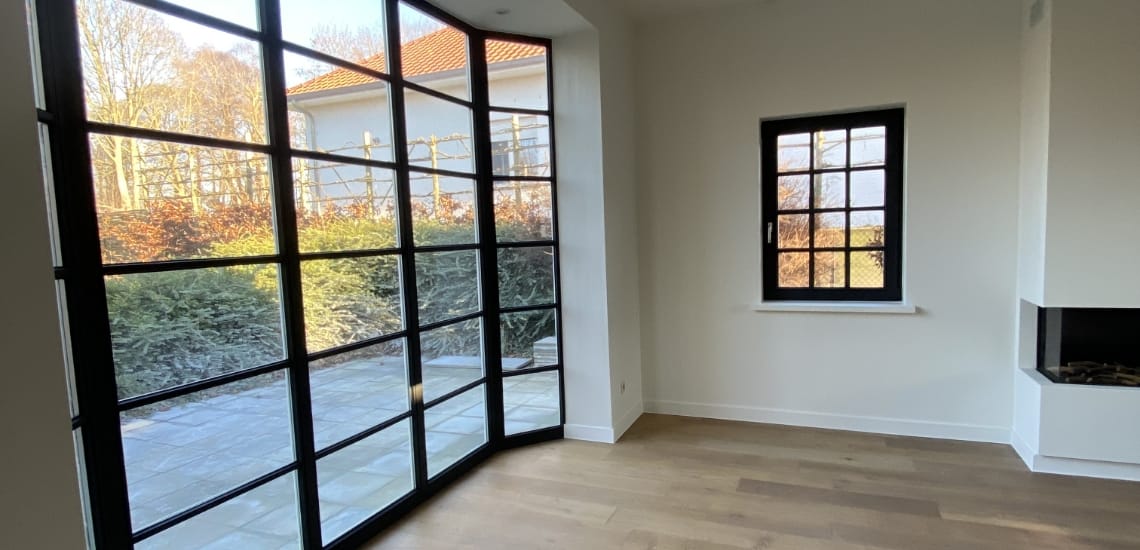 Een moderne kamer met grote zwarte ramen, lichte houten vloeren en een minimalistisch interieur. Het uitzicht naar buiten toont groen en een naburig huis onder een heldere hemel, wat doet denken aan de elegante eenvoud van de restauratie van een boerderij.