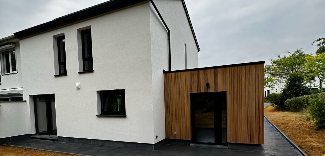 Een modern huis heeft een gerenoveerd wit gedeelte van twee verdiepingen naast een gedeelte van één verdieping met houten lambrisering. Het gebouw, gemarkeerd door zwarte raamkozijnen, staat tegen een bewolkte lucht. Een geplaveide oprit leidt ernaartoe, met weelderig groen dat de achtergrondambiance versterkt.
